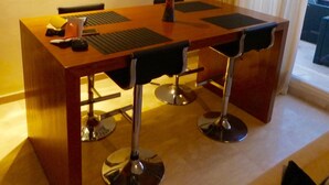 Uma área de refeições moderna com mesa de madeira e banquetas, um detalhe decorativo na parede e vista para o exterior através de uma janela com cortinas.