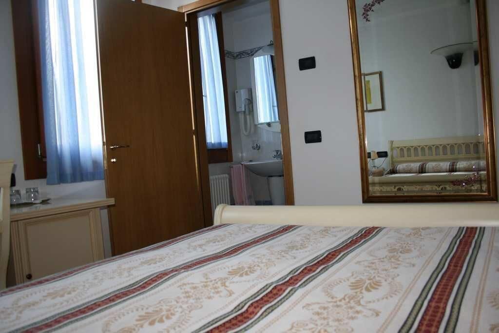 A bedroom with a bed, a wooden door, a mirror, and a bathroom visible through an open door.