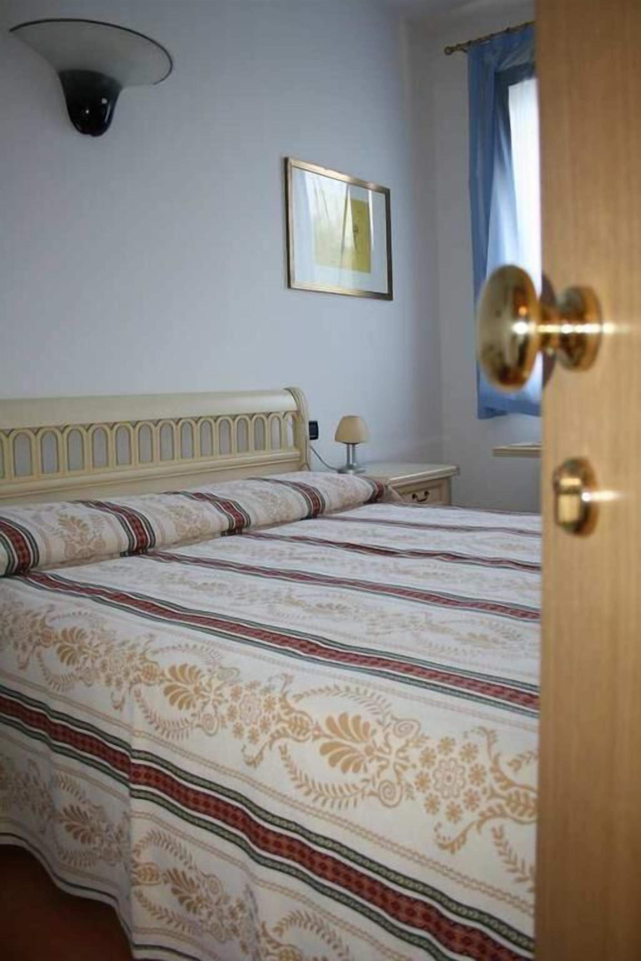 A bedroom with a bed, a bedside table, a lamp, a framed picture on the wall, and a door.