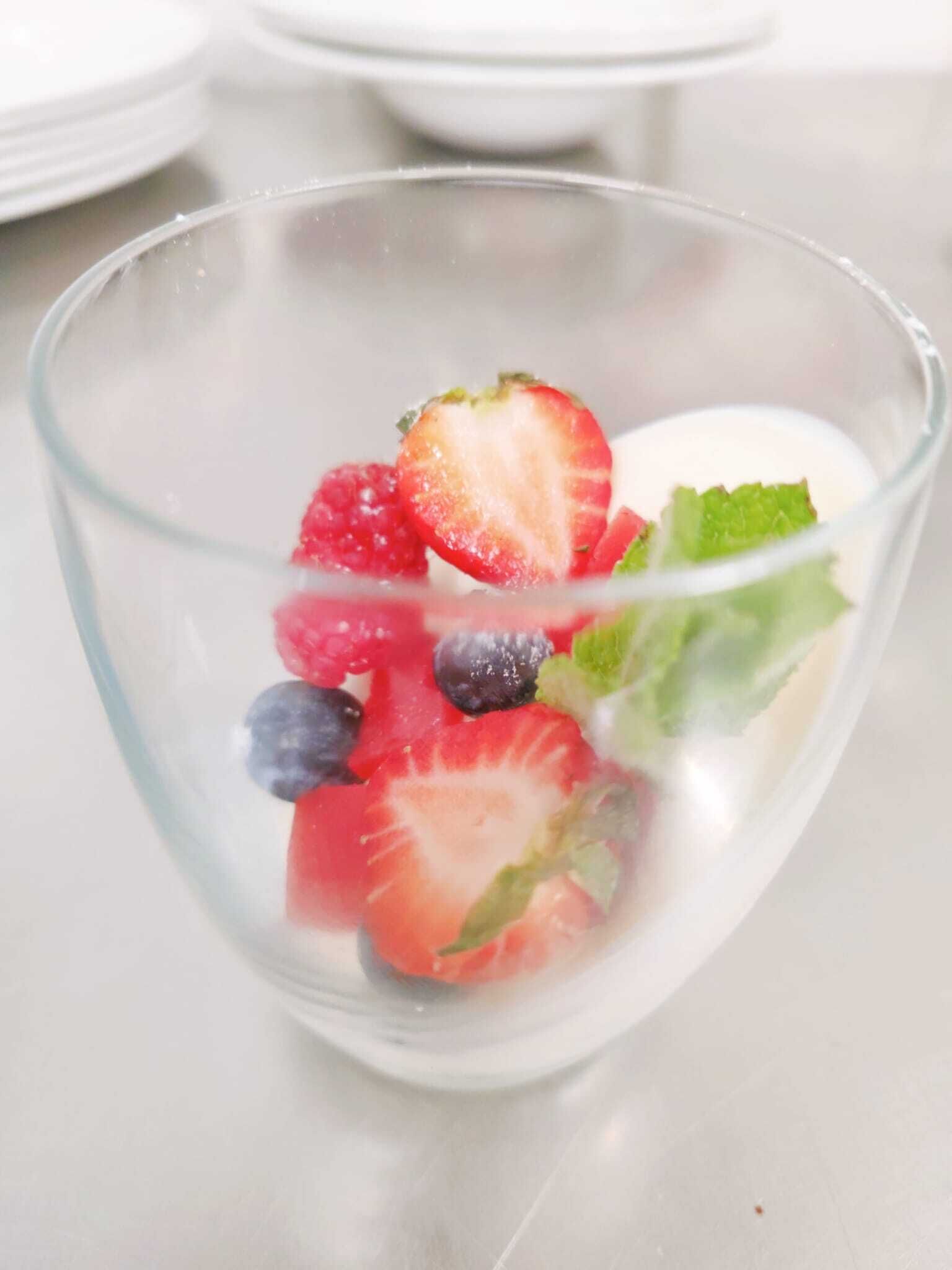 Un saladier en verre transparent contenant des fraises coupées en tranches, des myrtilles, des framboises et une feuille de menthe.