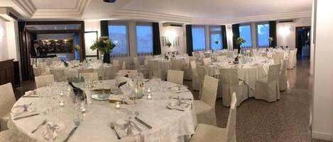 A banquet hall with round tables set for a formal event, featuring white tablecloths and chairs, and large windows with curtains.
