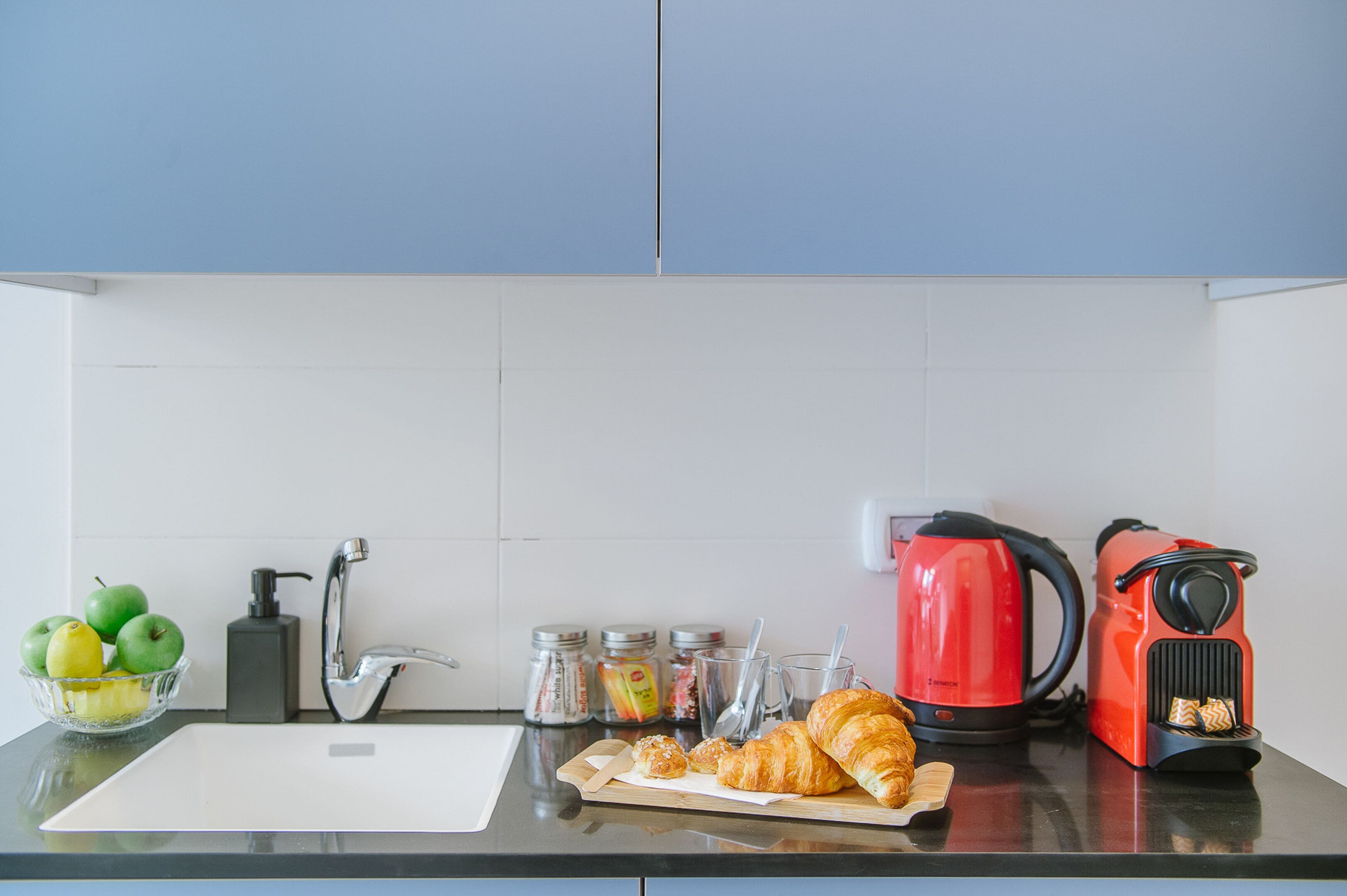 Un plan de travail de cuisine avec un évier, une planche à découper avec des croissants, une bouilloire rouge et des pots contenant des condiments.