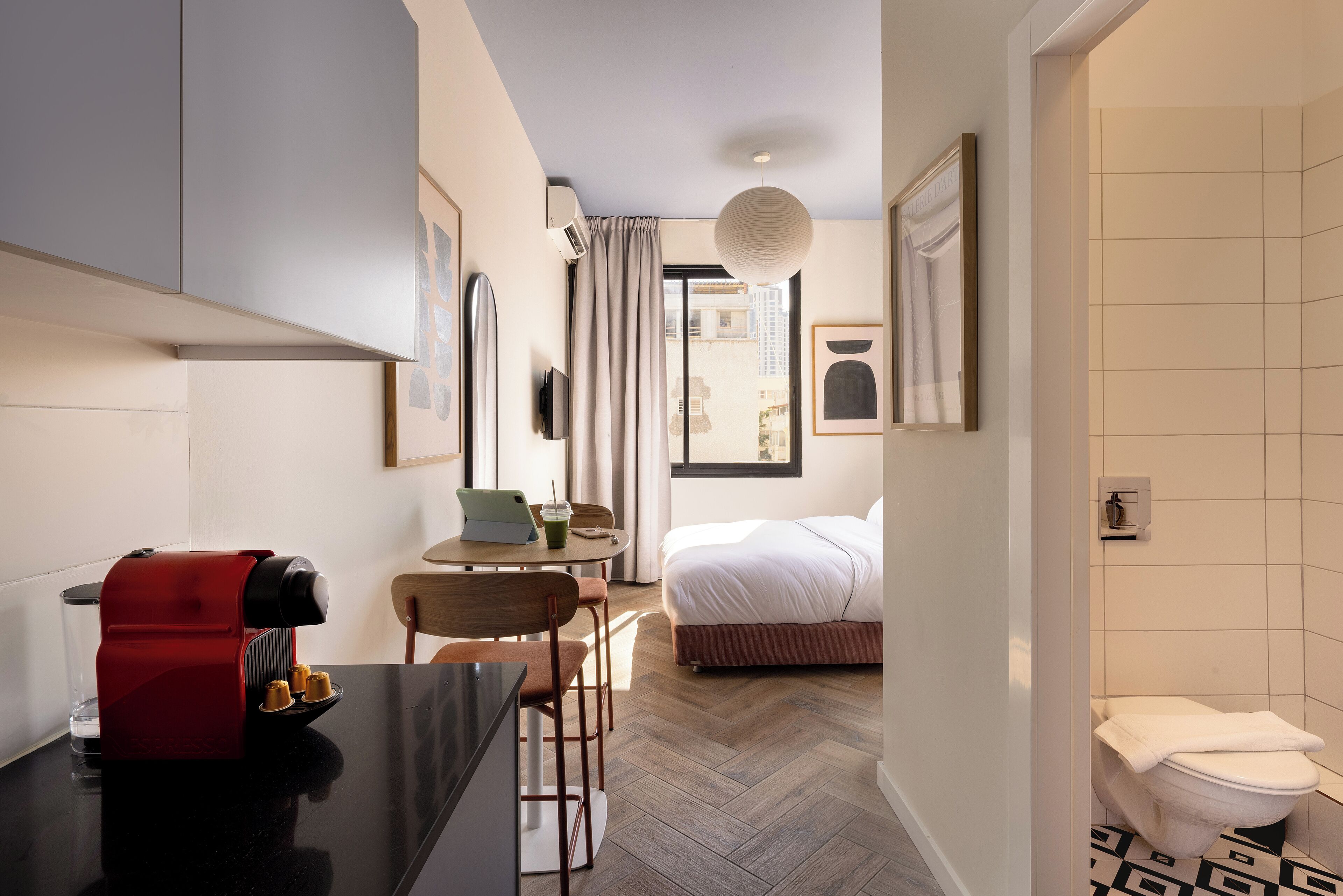 A hotel room with a bed, a desk with a coffee machine, a chair, a table with a laptop and a drink, and a bathroom with a toilet and a sink.