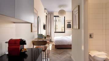 A hotel room with a bed, a desk with a coffee machine, a chair, a table with a laptop and a drink, and a bathroom with a toilet and a sink.