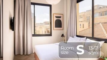 A hotel room with a bed, curtains, a window with a view of buildings, a wall-mounted air conditioner, and a framed picture.