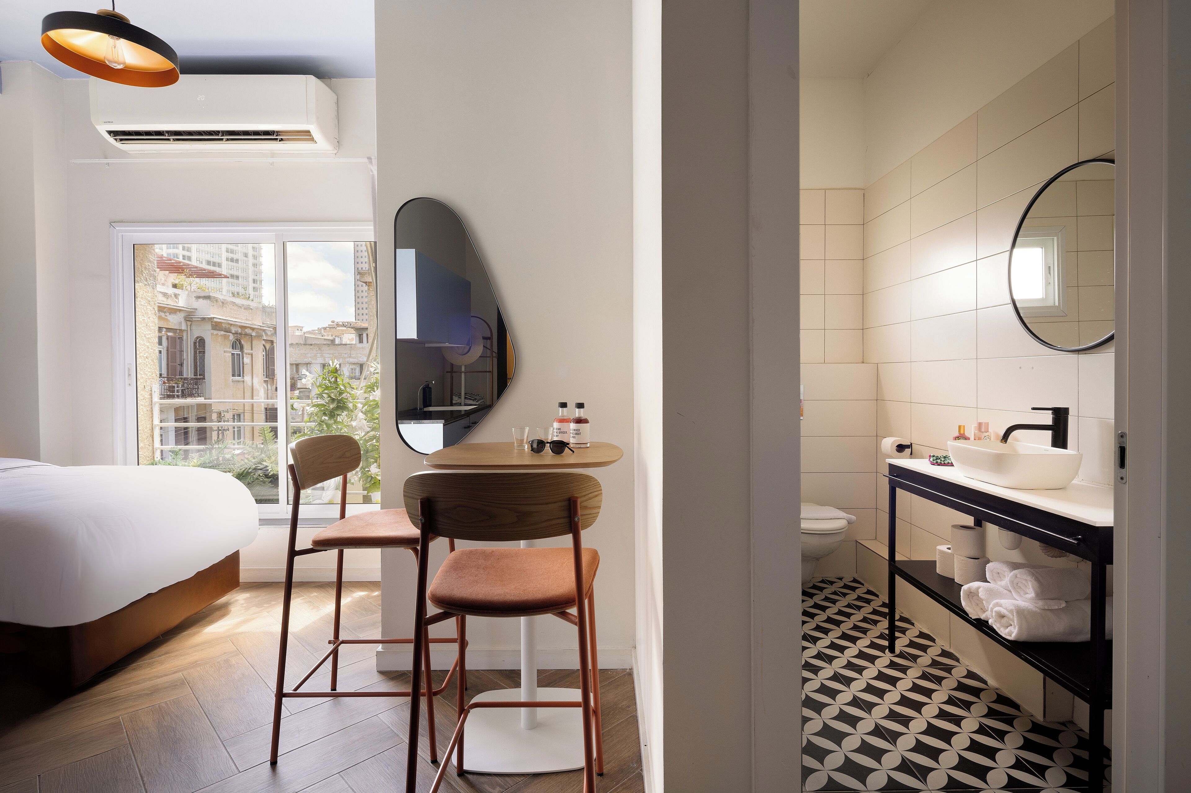 A modern hotel room with a bed, a small round table with chairs, a bathroom with a sink and mirror, and a view of buildings outside the window.
