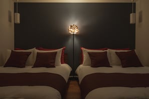 Hotelkamer met twee bedden, elk voorzien van wit beddengoed en rode accenten, een vloerlamp in het midden en een donkere muur.