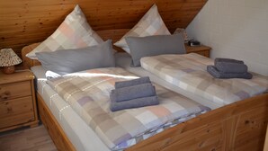 Ein schlicht eingerichtetes Schlafzimmer im Blockhausstil mit zwei Betten, jeweils mit kariertem Bettbezug und vielen Kissen. Ein hölzerner Nachttisch mit Lampe und ein kleiner Tisch mit einem Radio ergänzen das Zimmer.
