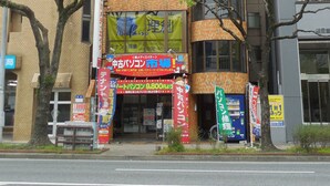 A building with a unique cylindrical section and multiple windows. There are several signs and banners in front, indicating various businesses. - AMP FLAT Nishijin 2F (Fukuoka)