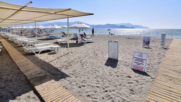 A beach with sun loungers and umbrellas, a wooden boardwalk, and a clear view of the sea and mountains in the background. - SUNSET BEACH APART (Fethiye)