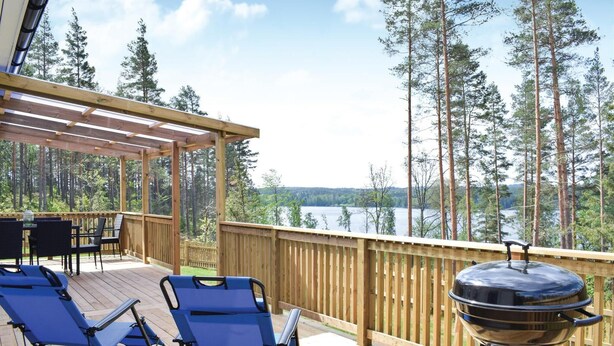 A wooden deck with two blue lounge chairs and a barbecue grill, overlooking a lake with tall trees.