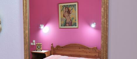 A bedroom with a pink wall, a wooden bed, a nightstand with a clock, and a framed picture above the bed.