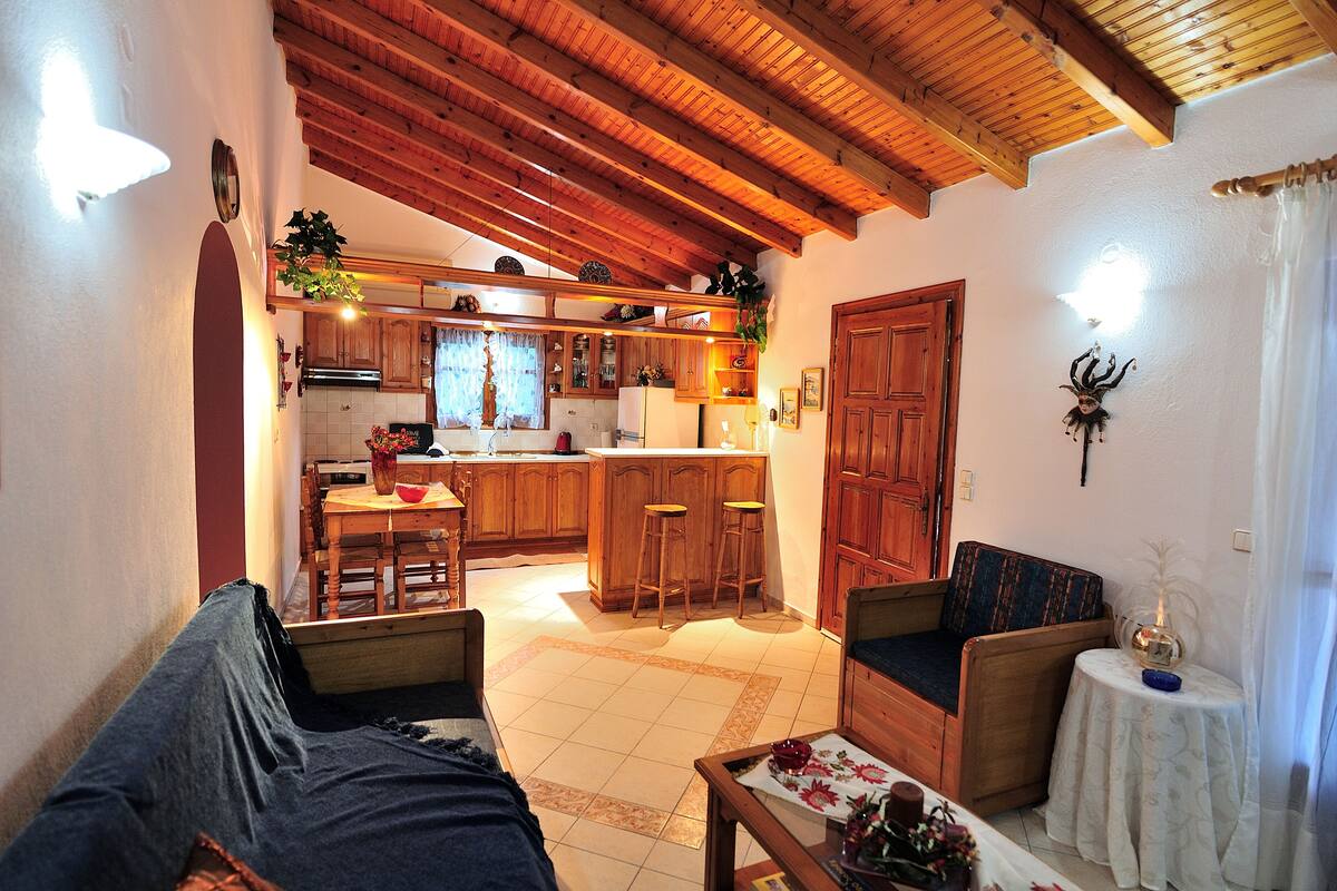 A cozy living room with a sofa, a coffee table, and a small table with a vase. There is a kitchen area with wooden cabinets and a dining table.