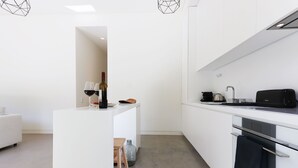 A modern kitchen with white cabinetry, a built-in oven, a coffee machine, and a small seating area with stools.