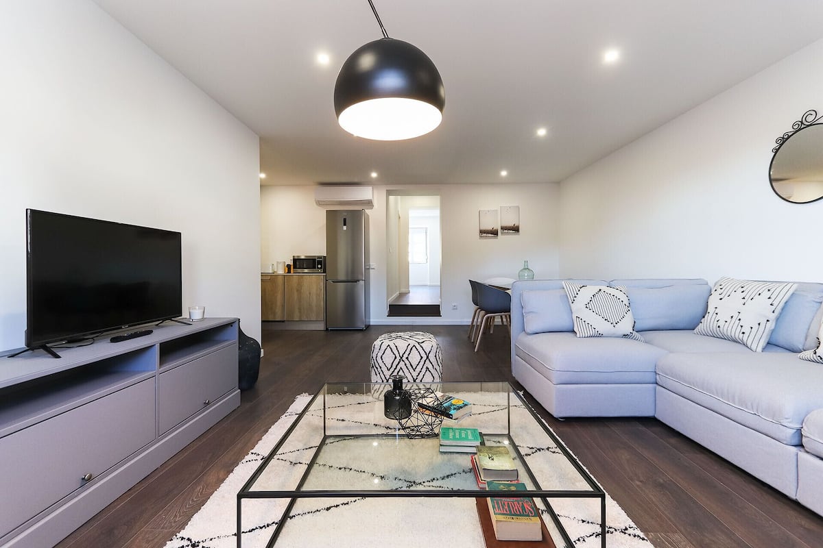 A modern living room with a flat-screen TV, a glass coffee table, a light blue sofa, and a kitchen in the background.