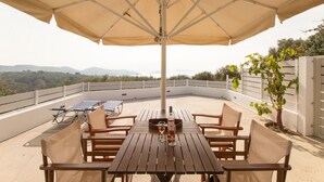 A wooden table with beige chairs and a large beige umbrella, set on a rooftop terrace with a view of distant mountains. - Armamento Villa (Skiathos)