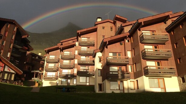 A modern multi-story building with balconies, set against a backdrop of a mountain and a rainbow.