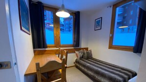 A compact room with a wooden desk, a cushioned bench, and a bed with a fur-like cover. There are two windows with curtains, and a picture frame on the wall.