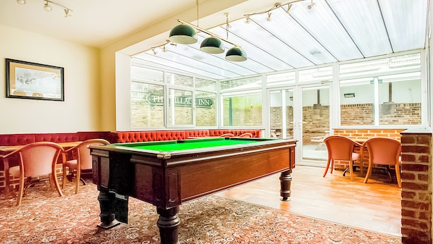 A pool table in a room with a patterned carpet, a framed picture on the wall, and a seating area with chairs.