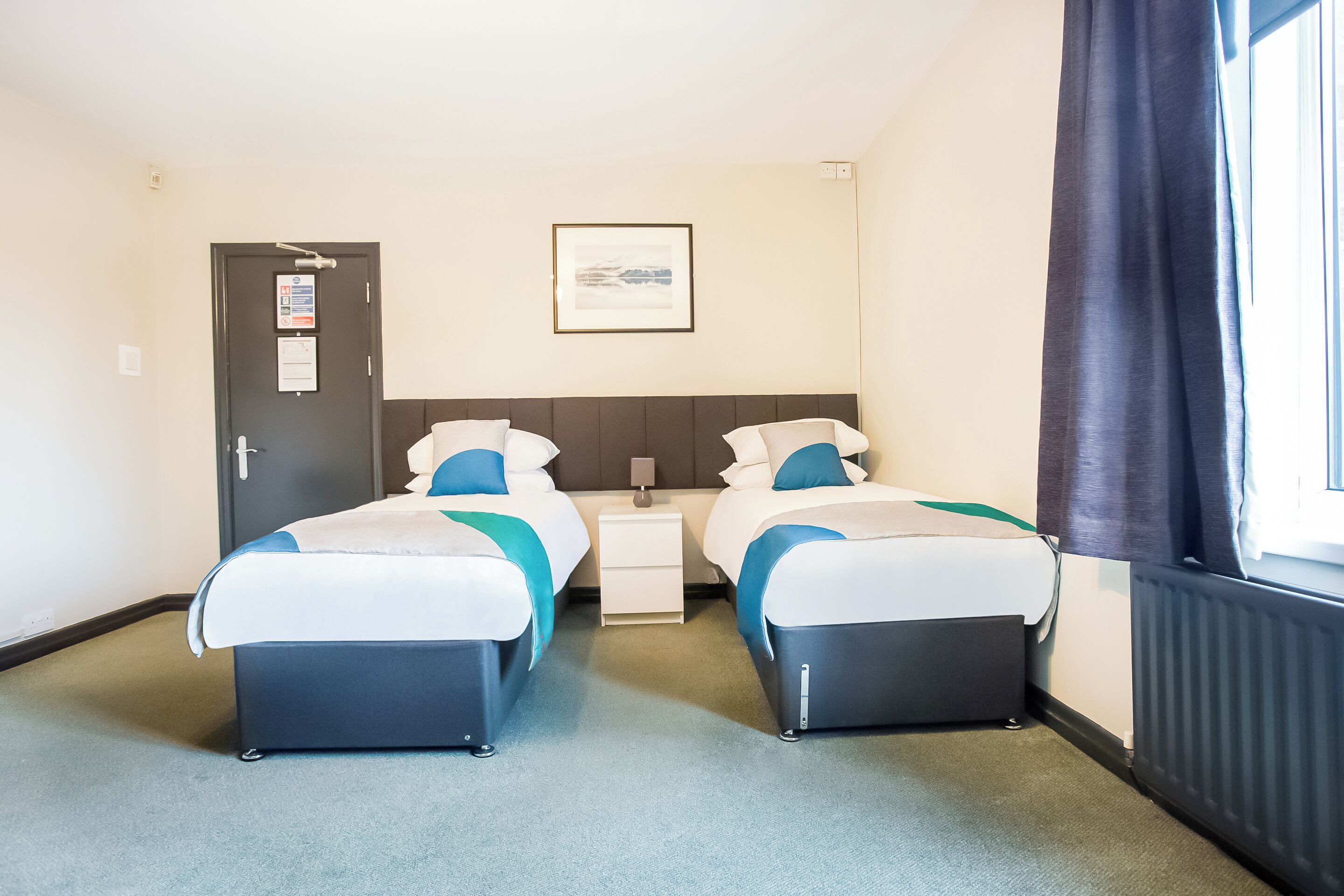 Habitación de hotel con dos camas, una mesita de noche pequeña, un cuadro enmarcado en la pared y una ventana con cortinas.