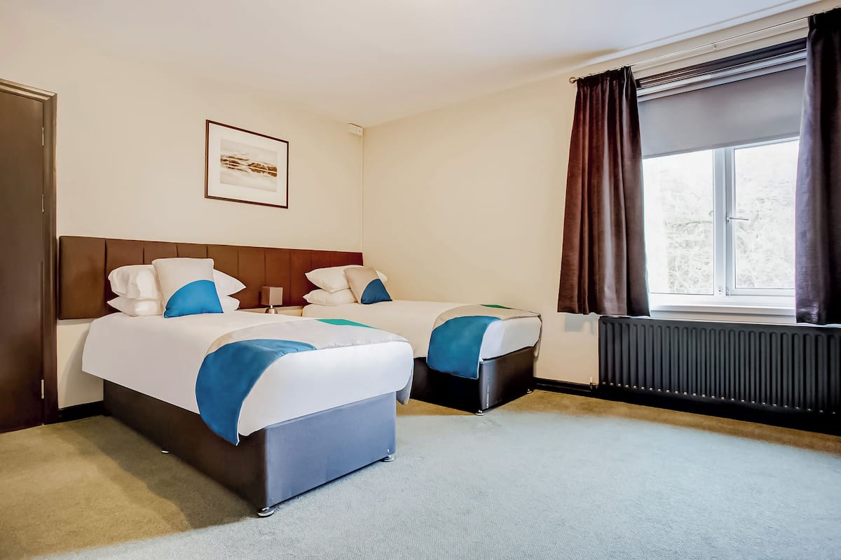 A hotel room with two beds, a framed picture on the wall, a window with curtains, and a radiator.