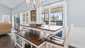 A dining area with a wooden table, white chairs, a chandelier, and a view of buildings through large windows. - Las Palmas: Spacious 4BR, 2BA home in Holden Beach, ideal for family getaways. (Holden Beach)