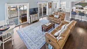 A spacious living room with a large leather sofa, a coffee table, and a TV. There is a dining area with a table and chairs, and a kitchen area with bar stools. - Las Palmas: Spacious 4BR, 2BA home in Holden Beach, ideal for family getaways. (Holden Beach)