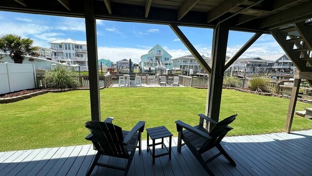 A covered deck with two Adirondack chairs and a small table, overlooking a grassy yard with a view of houses in the background. - Las Palmas: Spacious 4BR, 2BA home in Holden Beach, ideal for family getaways. (Holden Beach)