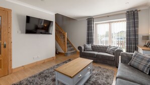 A living room with a grey sofa, a wooden coffee table, a TV mounted on the wall, a staircase, and a window with curtains.