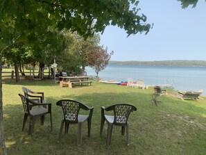 A lakeside picnic area with wooden benches and plastic chairs, a grill, and a dock extending into the water.