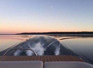 A boat's wake on calm water with a clear sky and a crescent moon.