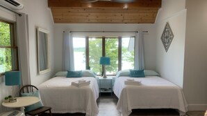 A hotel room with two beds, a small table, a chair, and a window with a view of greenery.