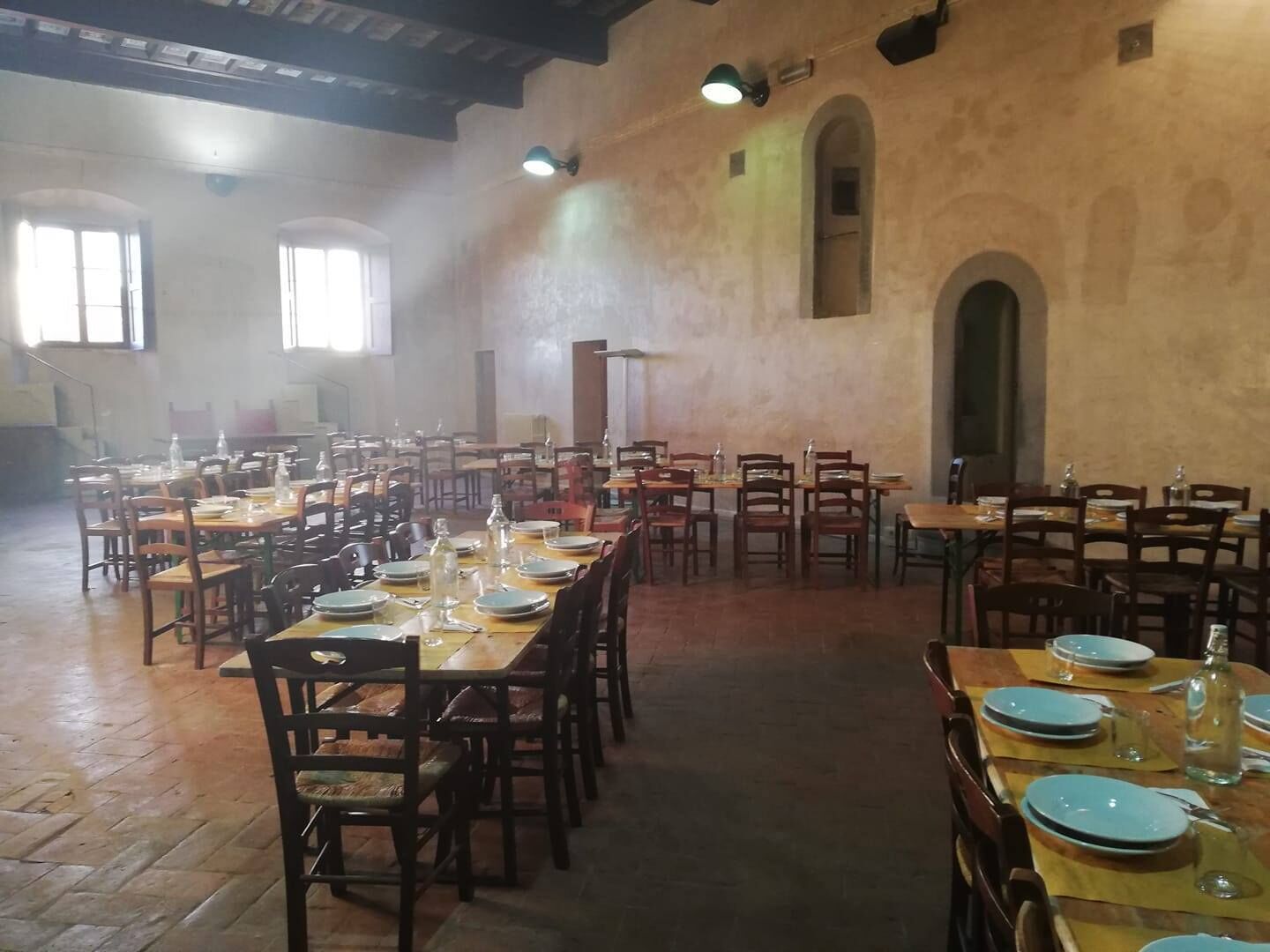 A spacious dining hall with multiple tables set for meals, wooden chairs, and a high ceiling with exposed beams.