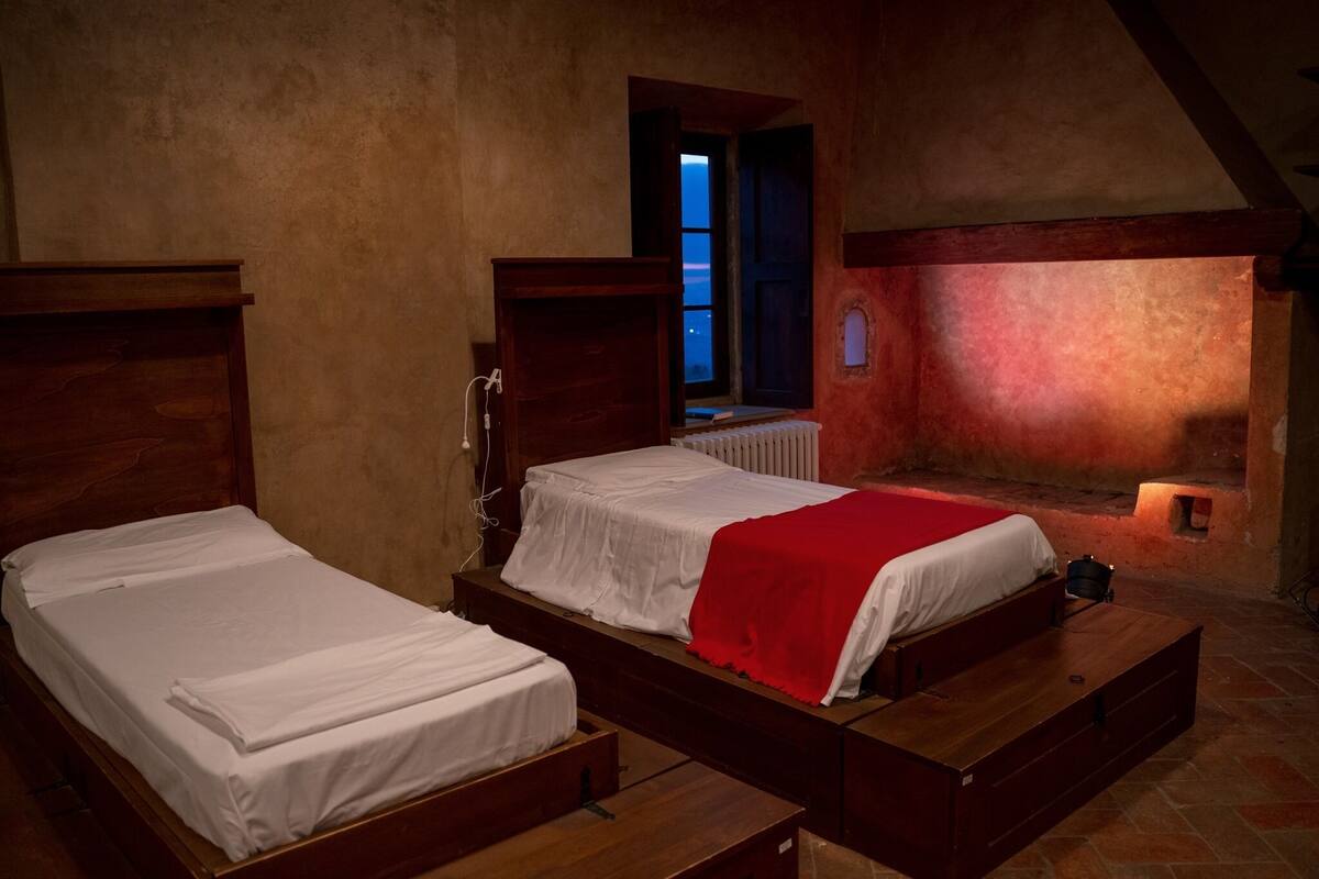 A room with two wooden beds, a red blanket on one, and a window with a view of the outside.
