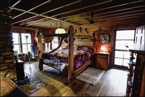 Een eenvoudige slaapkamer in een houten blokhut, met een houten bed, een bureau en een groot raam.