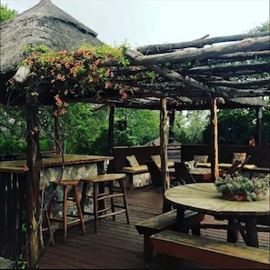 Een terras van hout buiten, met rustieke meubels, een rieten constructie en een pergola begroeid met klimplanten.