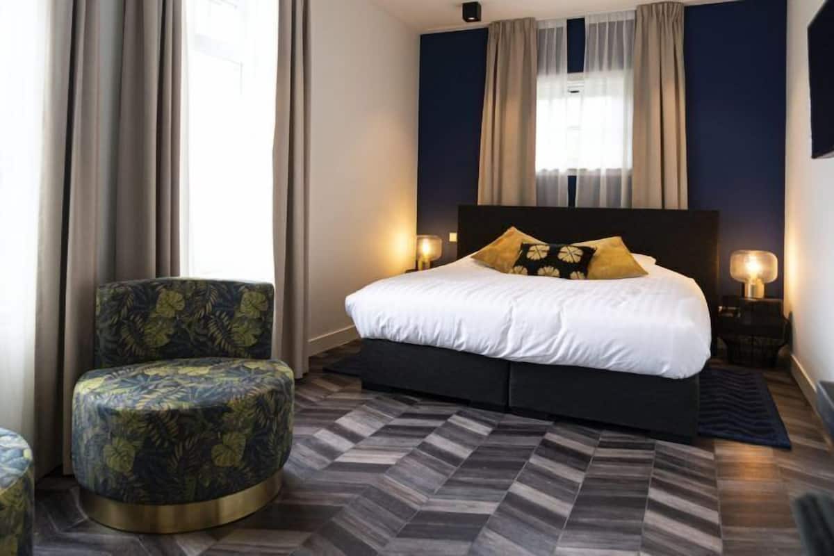A modern hotel room with a large bed, a round armchair, and a patterned ottoman.