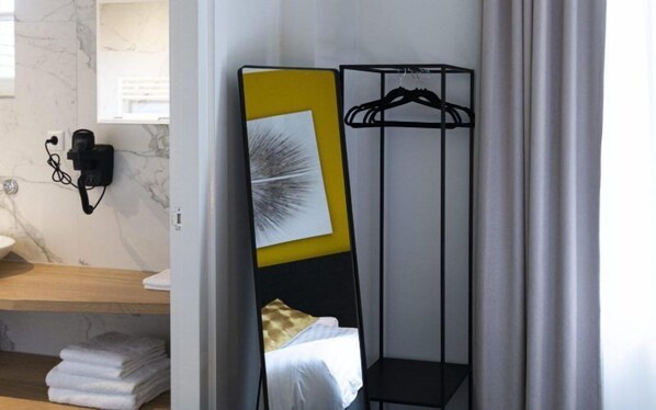 A compact hotel room with a bed, a bedside table, a wall-mounted telephone, and a black metal rack with a hanging garment.