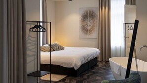 A modern bedroom with a bed, a bedside table, a hanging clothes rack, a large mirror, and a bathroom with a bathtub.