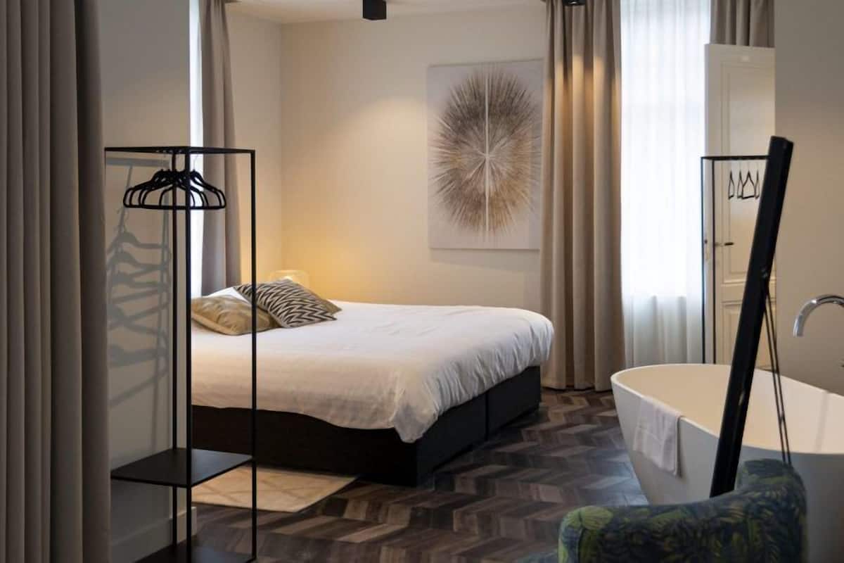 A modern bedroom with a bed, a bedside table, a hanging clothes rack, a large mirror, and a bathroom with a bathtub.