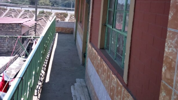 Un balcó estret amb un paviment de rajoles, baranes verdes i un mur de maó vermell.