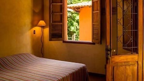Una habitación con una cama, una ventana con contraventanas abiertas, una lámpara y una puerta de madera con un panel de vidrio decorativo.