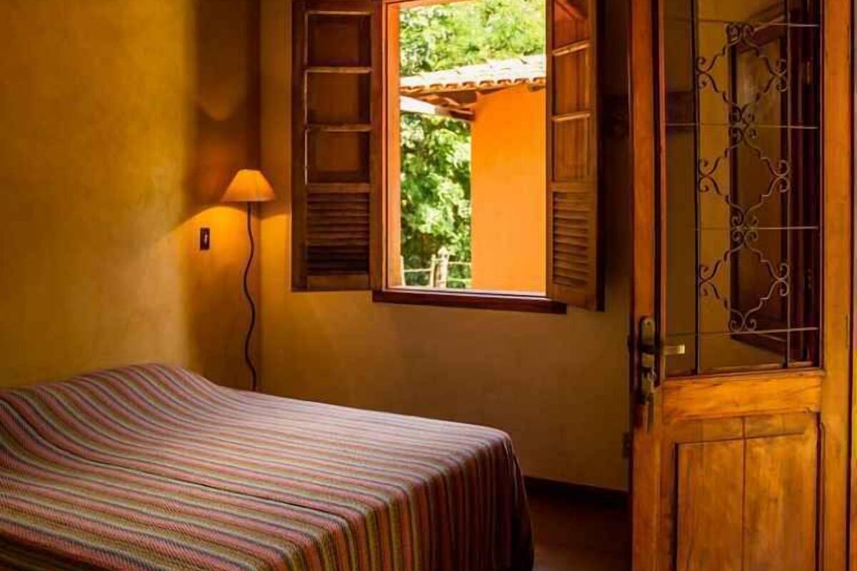 Una habitación con una cama, una ventana con contraventanas abiertas, una lámpara y una puerta de madera con un panel de vidrio decorativo.