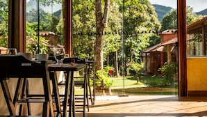 Comedor con mesa y sillas, una pared divisoria de vidrio y vista a un jardín y montañas.