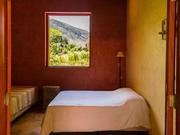 A bedroom with a bed, a window with a mountain view, a bedside lamp, and a small bedside table.