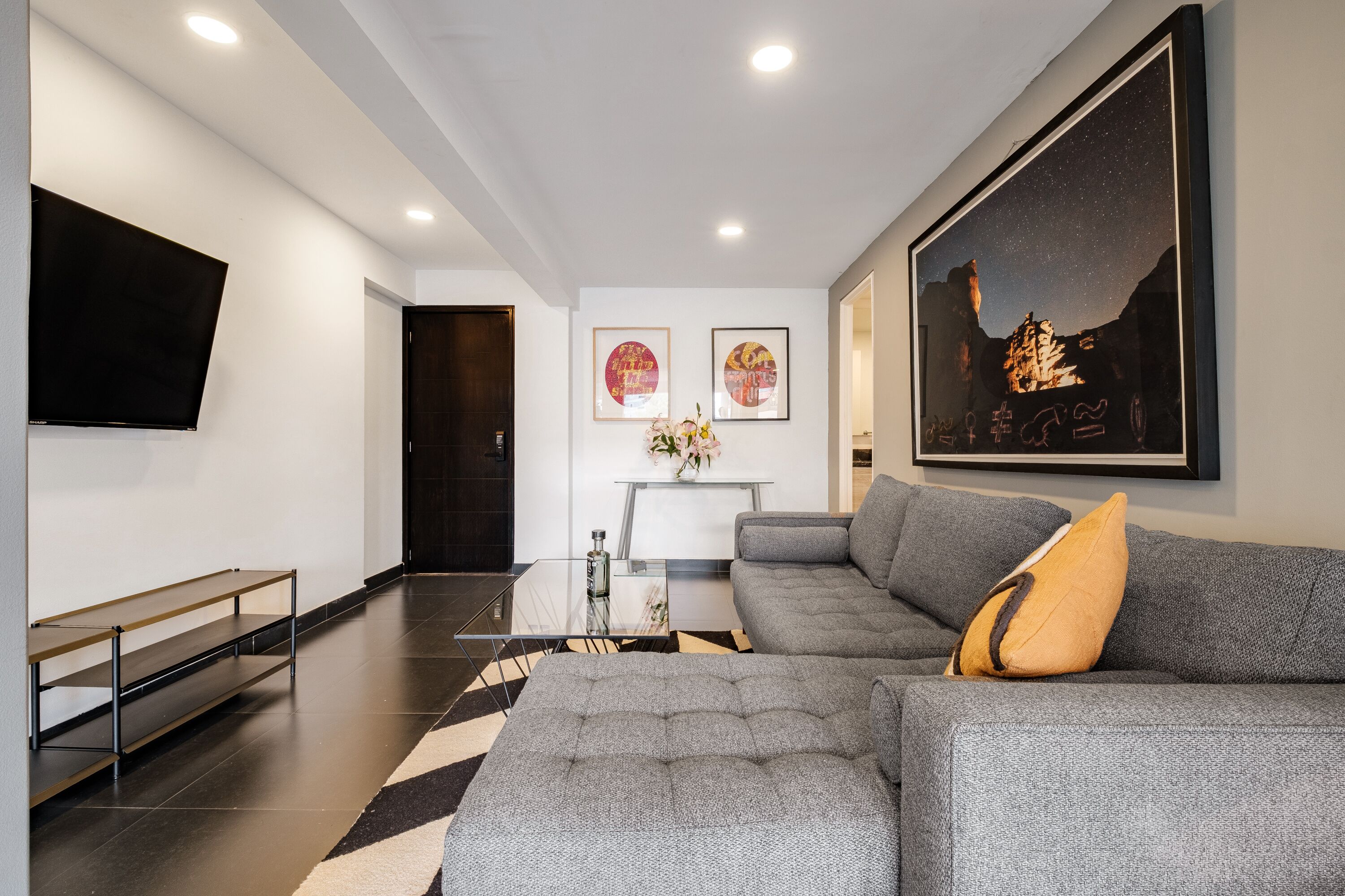 Un salon moderne avec un canapé gris, une table basse, un téléviseur à écran plat fixé au mur et des œuvres d’art accrochées aux murs.