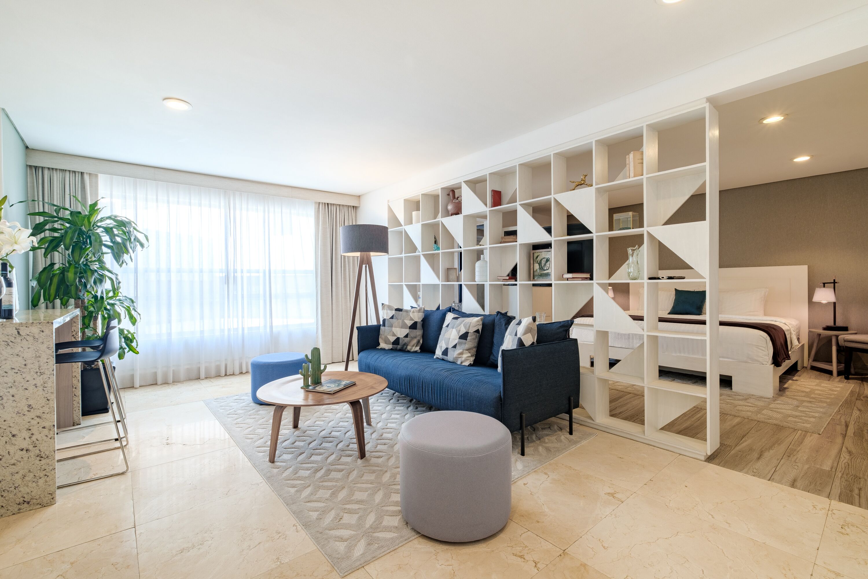 A modern living room with a sofa, coffee table, and ottoman. There is a bookshelf and a lamp.