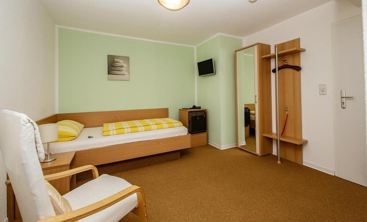 A single-bed room with a wooden headboard, a bedside table, a chair, a TV mounted on the wall, and a wardrobe with a mirror.