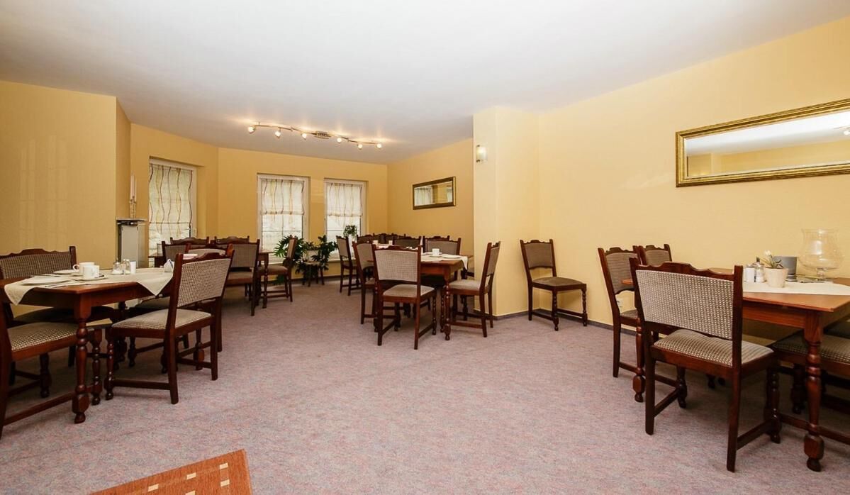 A hotel dining area with multiple wooden tables and chairs, a carpeted floor, and large windows with curtains.