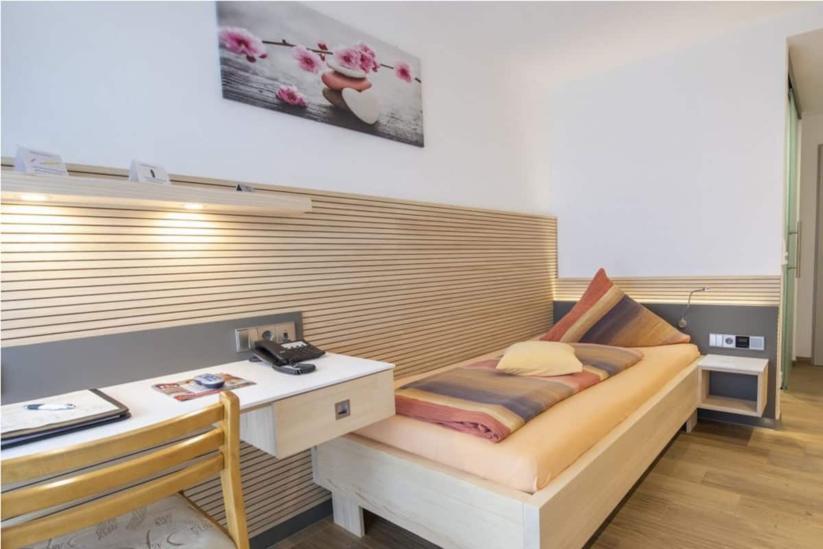 A modern hotel room with a wooden headboard, a desk with a telephone, a bed with pillows, and a painting on the wall.